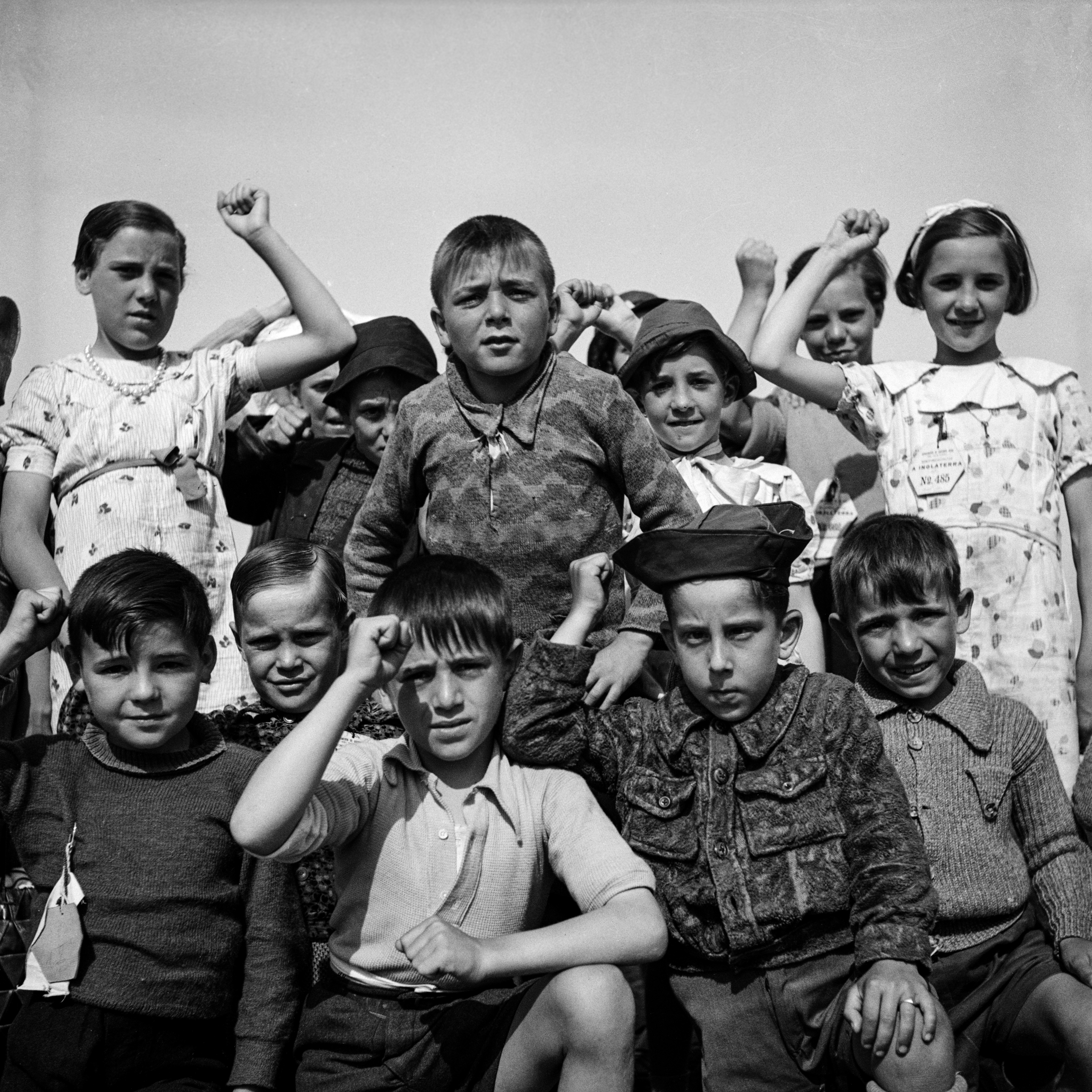 A group of around a dozen children of varying ages pose together outdoors, several raising clenched fists. Many wear numbered identity labels pinned to their clothing. Their expressions range from defiant to solemn. The image is square-format with a clear, even sky behind them.