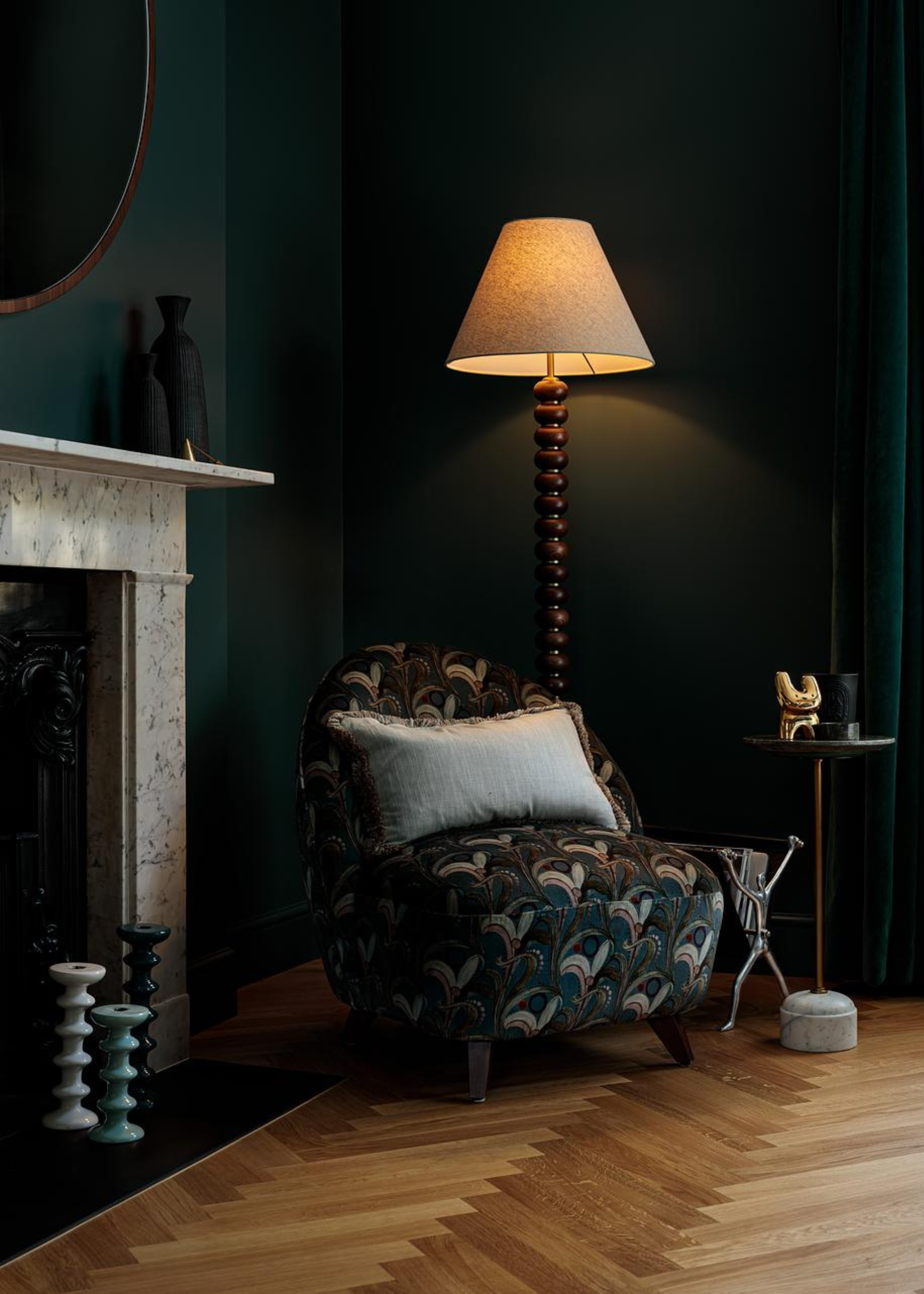 A dark green living room corner with a bobbin lamp near a patterned sofa seat, apredestal side table, a magazine holder and a set of candlesticks near a fireplace