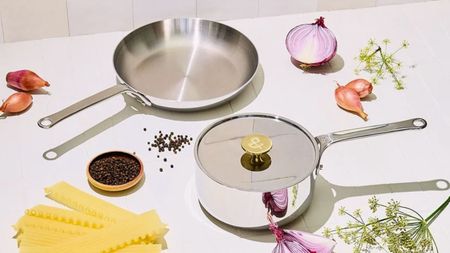 Two stainless steel pans on a white counter beside raw ingredients like onions, pasta, and herbs