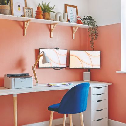 A coral-painted home office with a white desk and floating shelf and a blue velvet chair