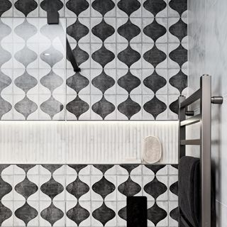 close up of a shower enclosure clad in patterned black and white tiles and pale marble tiles, with a recessed lit shelf, chrome towel rail and a glass screen