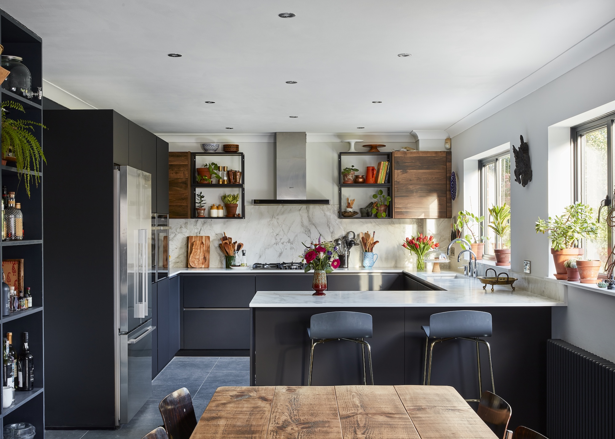 A u shaped kitchen in navy blue with a large island and bar stools beside it. There is open shelving on either side of the stove as well as an extractor fan. A view of the fridge is visible as well as the dining table