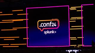 The Splunk .conf24 logo on a keynote screen live at Splunk .conf24 in Las Vegas. Decorative: The scene is lit in neon purple and orange lighting.