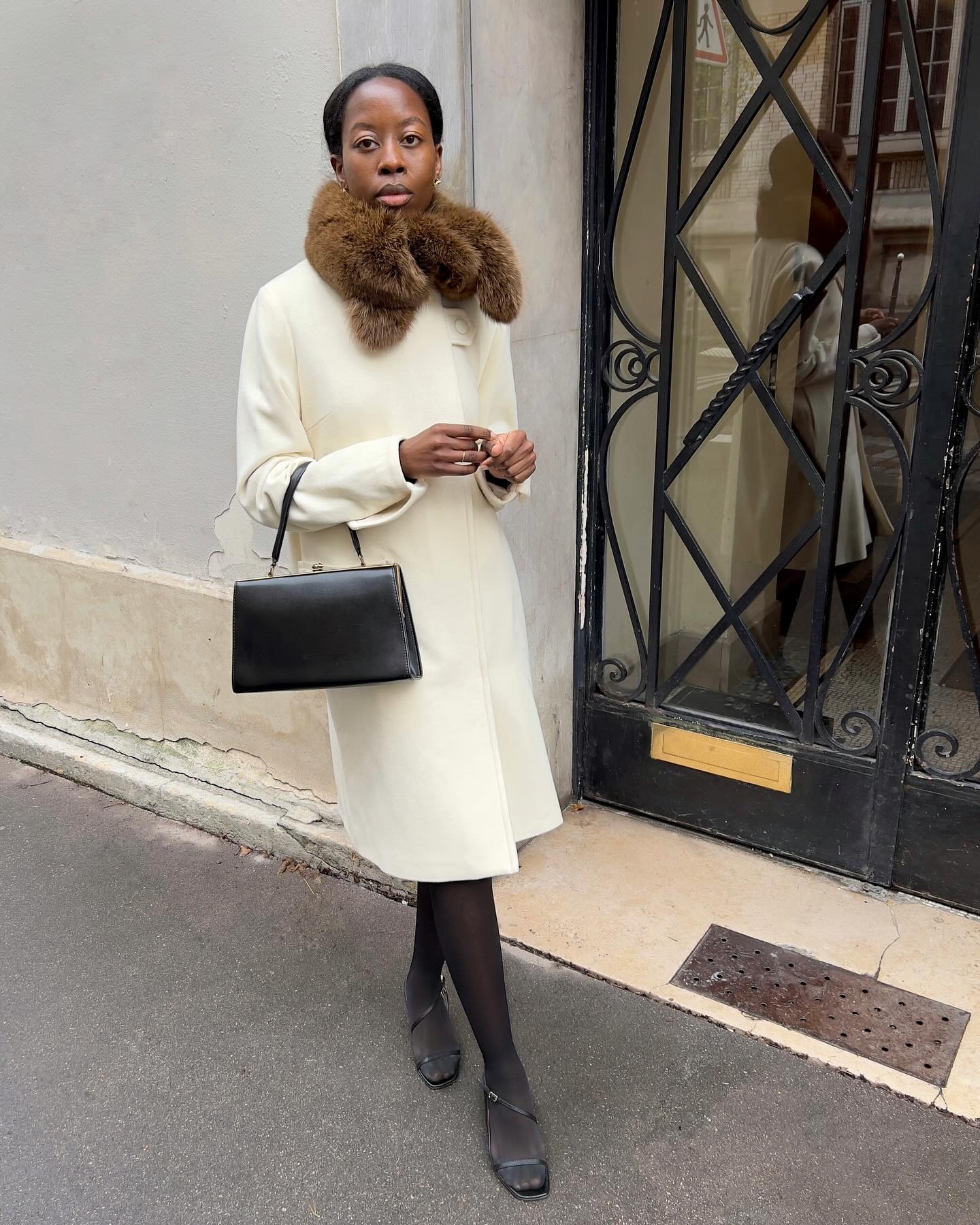 @sylviemus_ wears a white car coat, a fur scarf, a black leather bag, sheer tights, and heels.