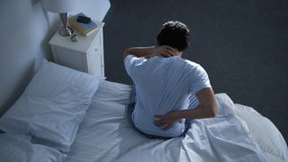 A man sits up in bed at night, holding one hand to his lower back because he&#039;s experiencing back pain