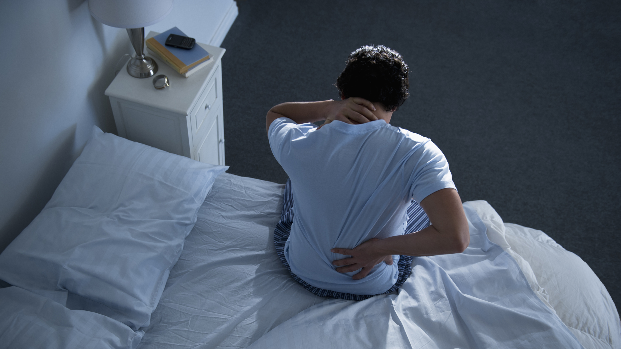 A man sits up in bed at night, holding one hand to his lower back because he's experiencing back pain