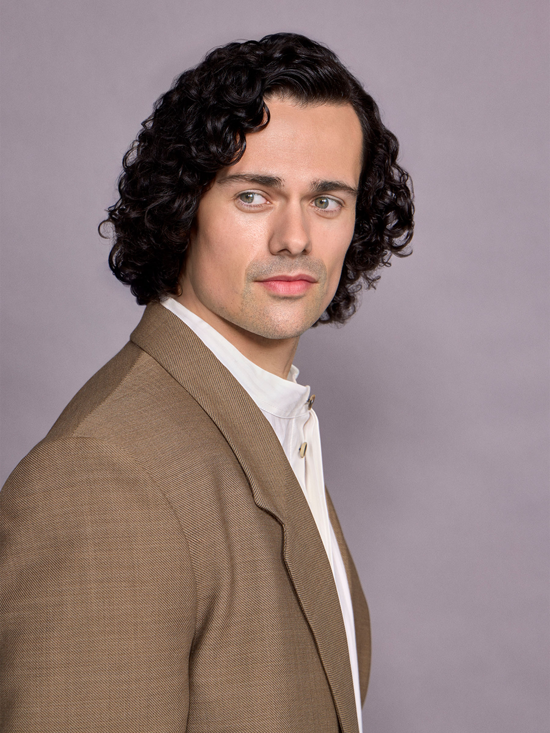 Actor Jamie Roy is looking off camera wearing a brown suit with a white tuxedo shirt.