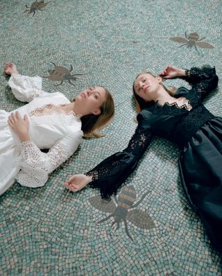 A woman in a lacy white dress, and another in a lacy black dress, lying on a mosaic-tiled floor