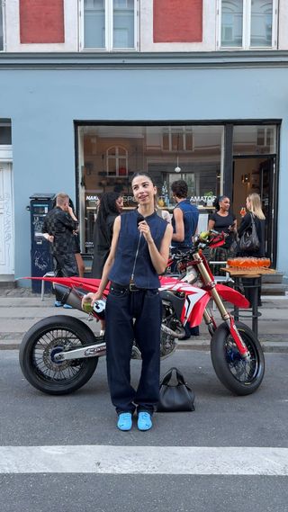 A woman wearing Puma Speedcat sneakers in blue with black trousers, a belt, and a navy vest.