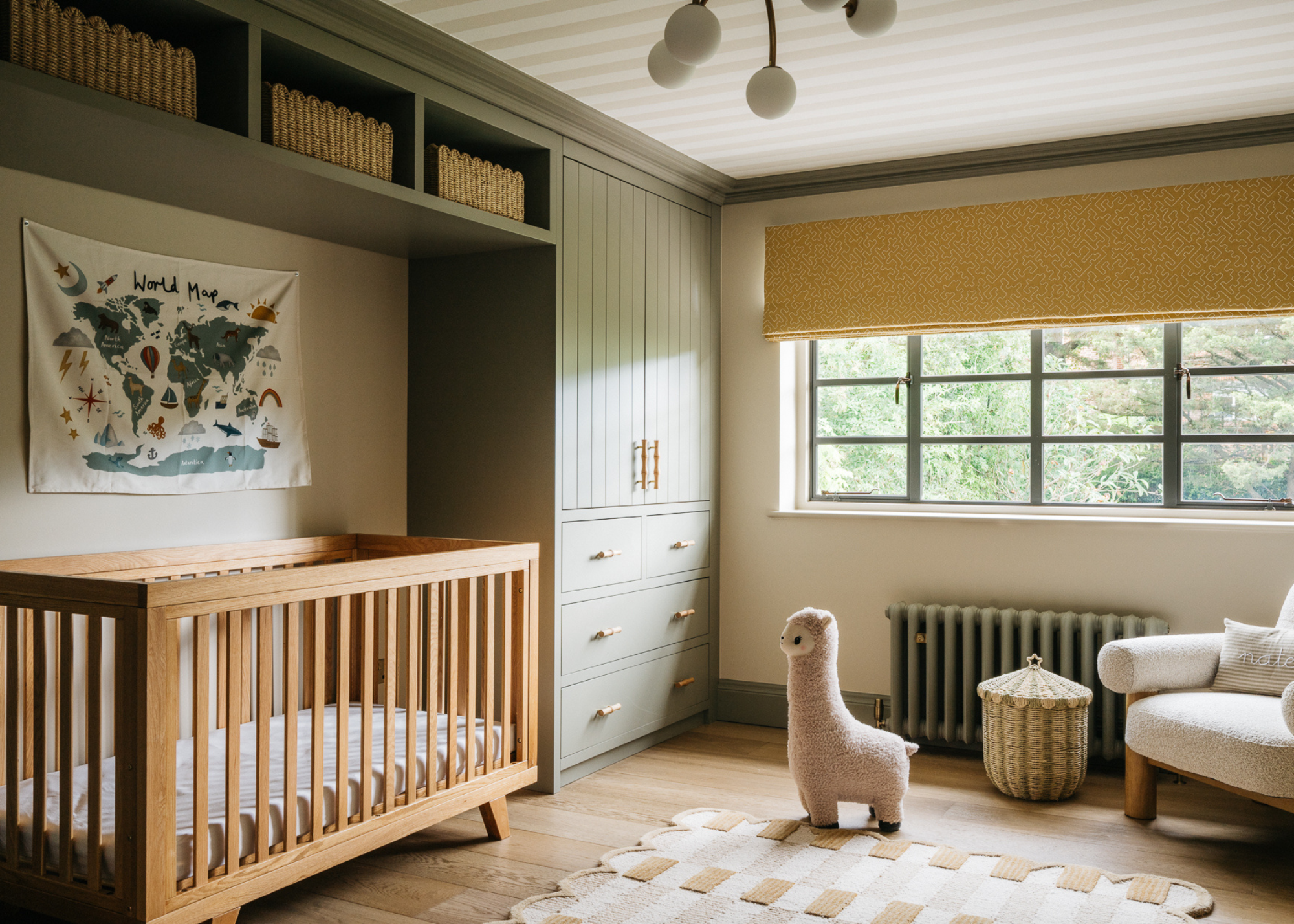 A nursery with a cot inset into wraparound built-in storage that's painted green, with wooden flooring, and a scalloped edge rug is in the center of the room with a toy llama on top and the window has a yellow roman blind with a white accent chair in the corner