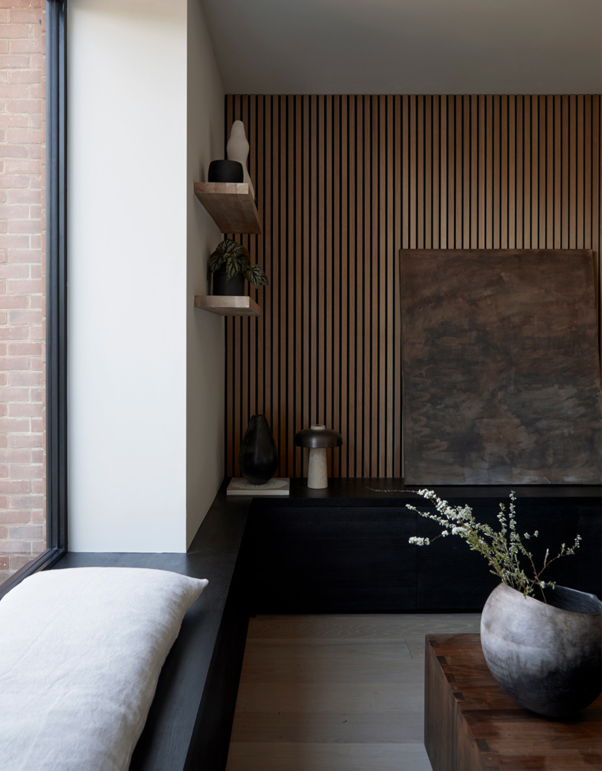 A window seat in the corner of an open plan living dining room with a wood panelled wall, a leaning painting, floating shelves, and a vase of stems