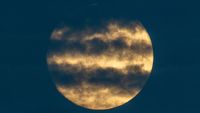 A yellow full moon glows in a dark sky with streaks of cloud lining its disk.