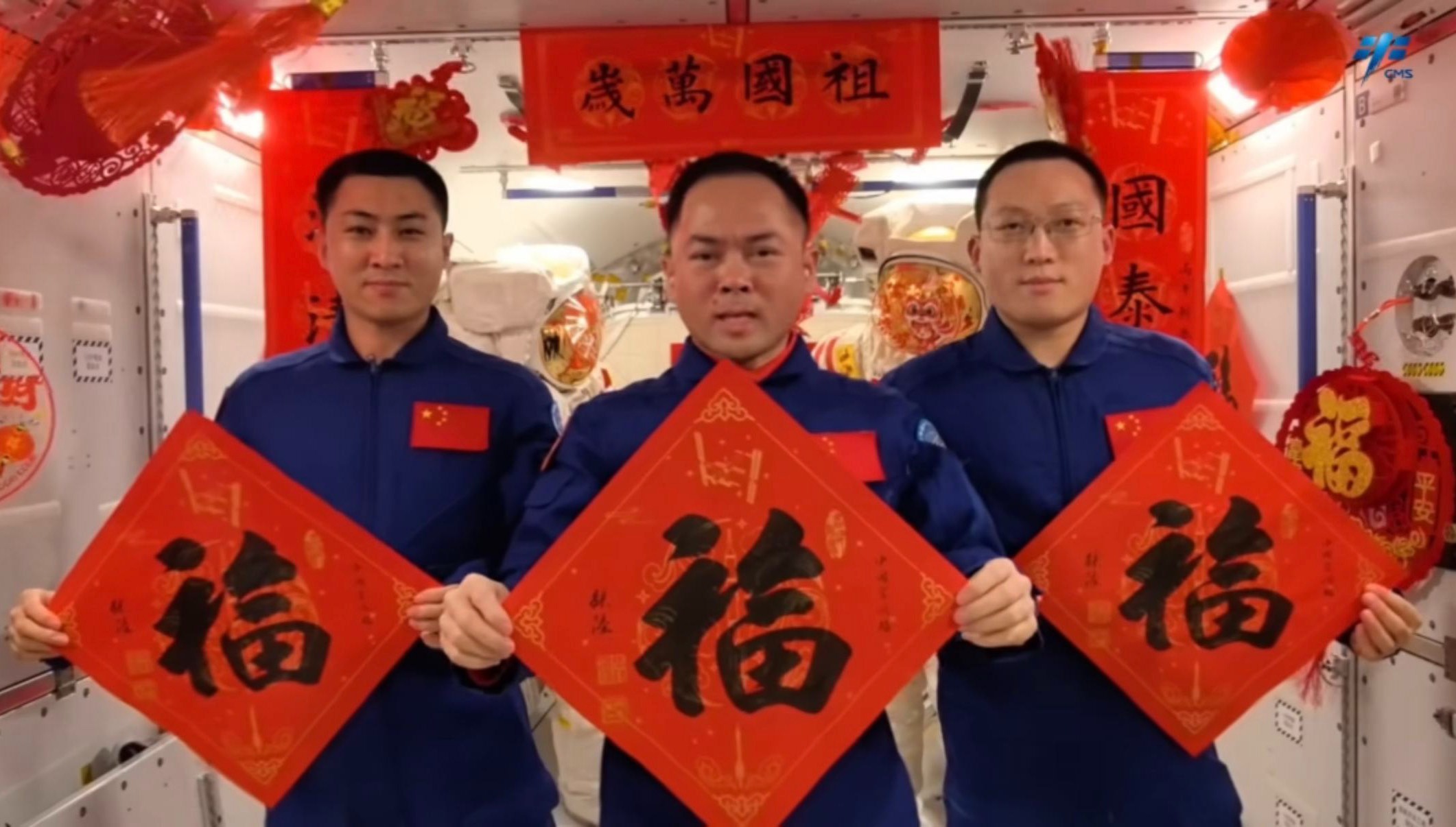 رواد فضاء يرتدون بدلات طيران زرقاء يقفون وسط زخارف حمراء وذهبية زاهية عليها الحرف الصيني 