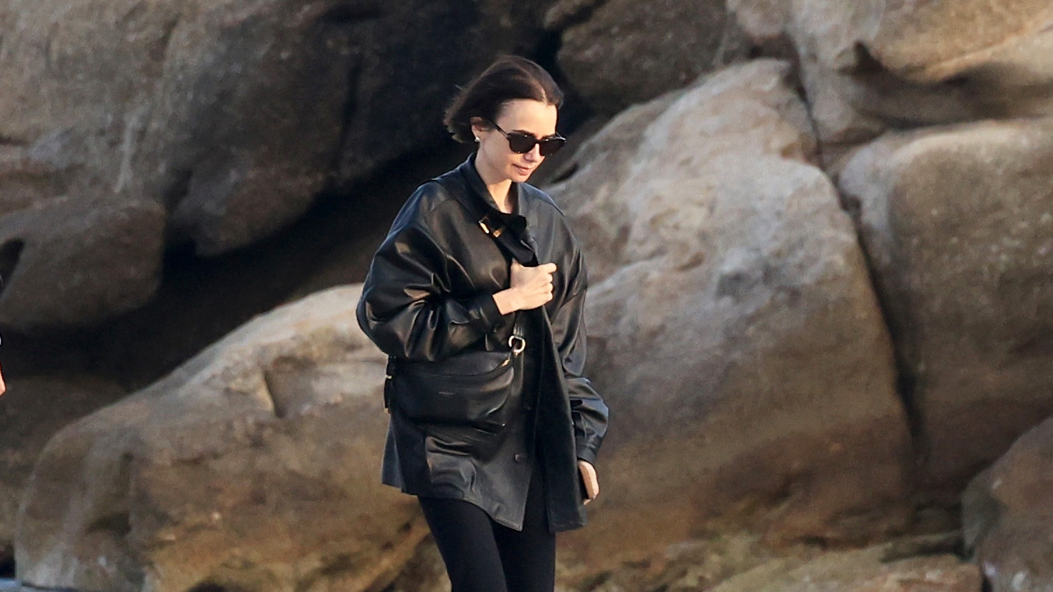 Lilly Collins wearing a black leather jacket, capri pants, and two-tone cream-and-black ballet flats in Mykonos