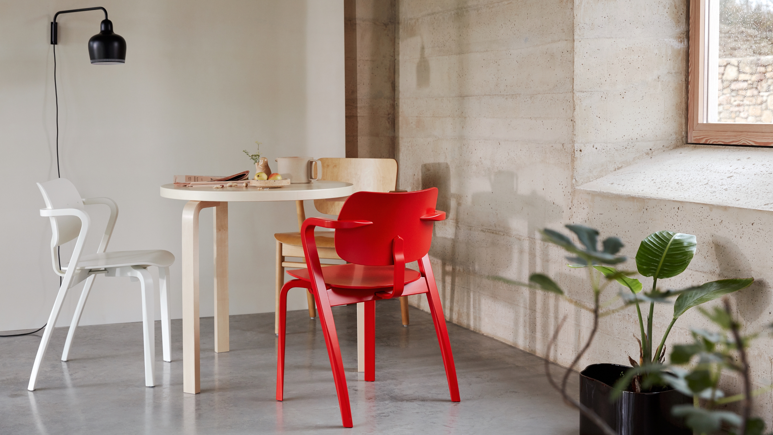 View of Scandinavian style, round dining table and chairs in white, wood and bright red. In the background is an industrial, concrete dining space.