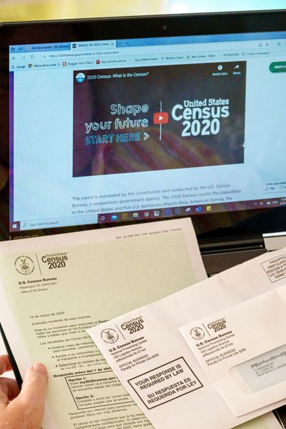 A hand holding, Census 2020, letter and forms, in front of computer displaying website