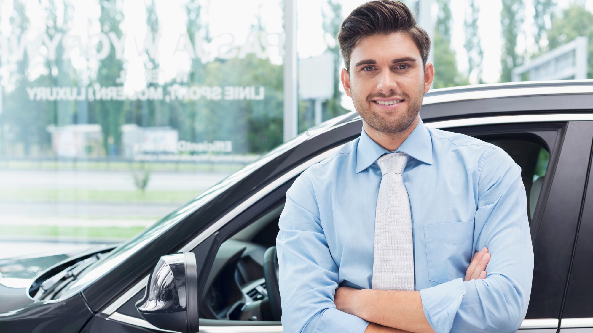 A friendly-looking, young male car salesperson stands in front of a car with his arms crossed.