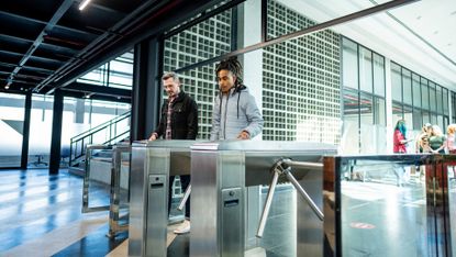 Two office workers walking into the lobby of their large, modern office building. Each is scanning their office card on their respective turnstile.