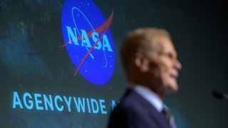 a man in a suit speaks into a microphone in front of a backdrop that reads "NASA"