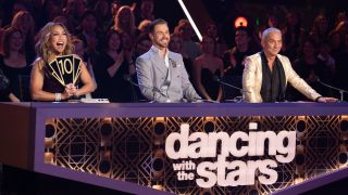 Carrie Ann Inaba holds up a 10 at the judges table while Derek Hough and Bruno Tonioli smile