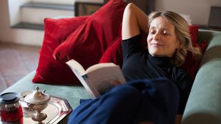 Woman lying down on sofa reading a book and drinking tea