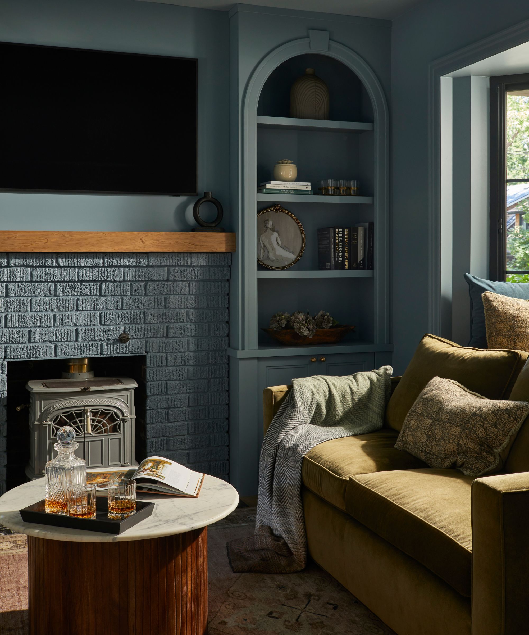 a blue color drenched living room with arched niche storage, a blue painted brick fireplace, green couch and a round marble topped coffee table