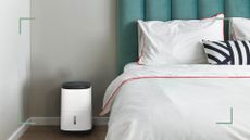 Meaco dehumidifier next to bed with white linen in a light grey bedroom to support an expert guide on What you need to know before buying a dehumidifier