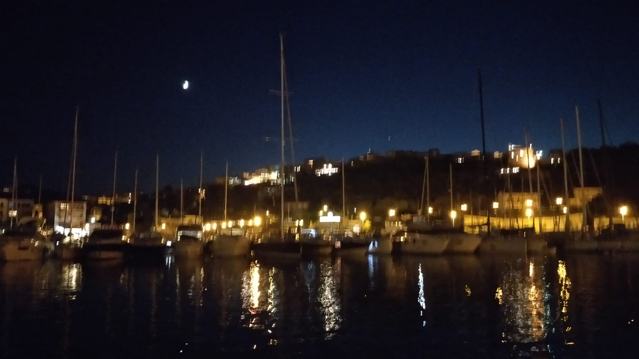 A moonlit marina full of sailboats reflects smeared streaks of golden light across the dark water.