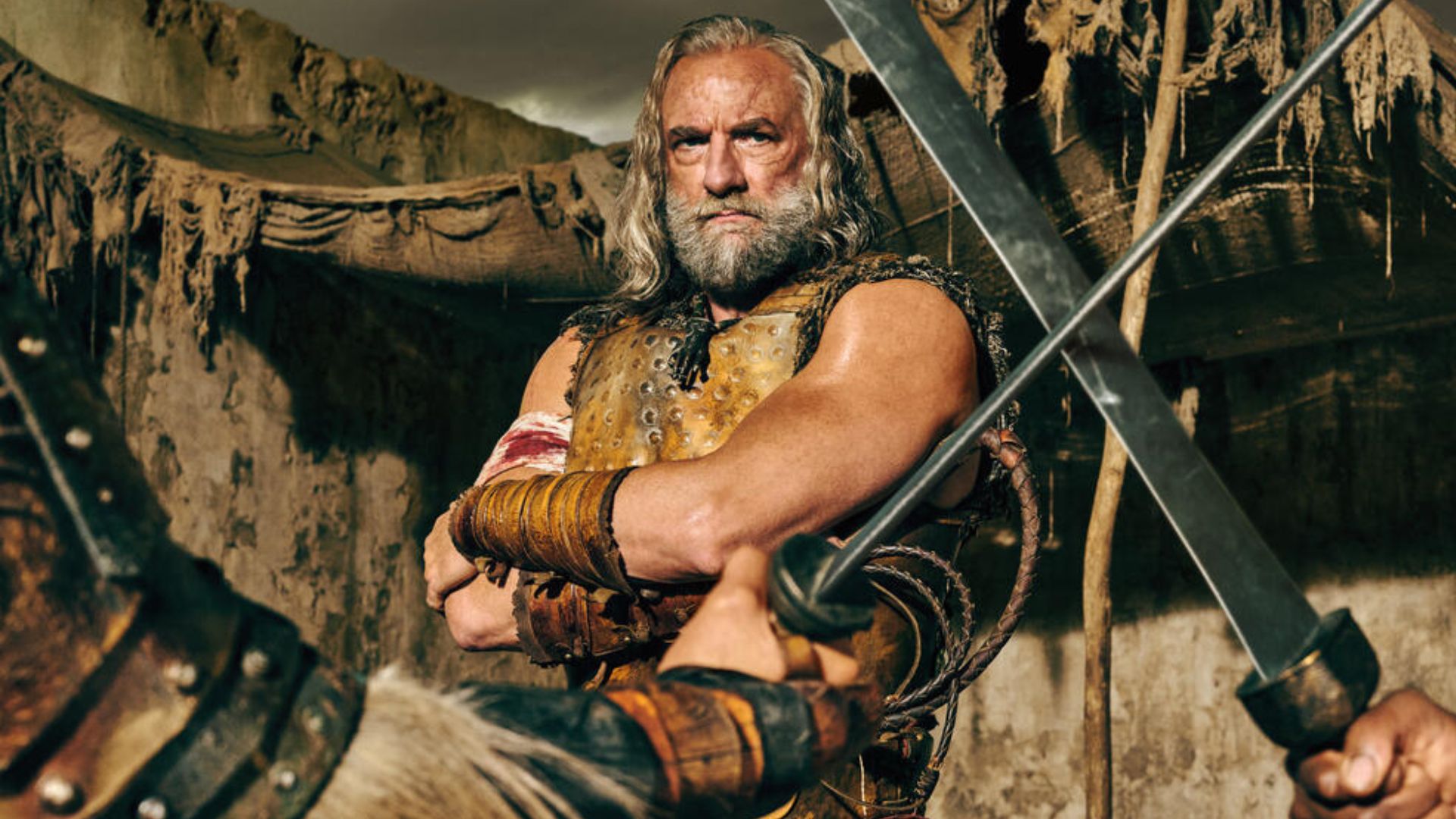 Korris stands with his arms folded watching two gladiators spar