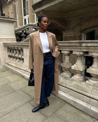 British fashion influencer Marilyn Nwawulor-Kazemaks posing on a London sidewalk wearing a winter outfit with a long camel coat, white top, black belt, dark-wash jeans, and round-toe flat black boots