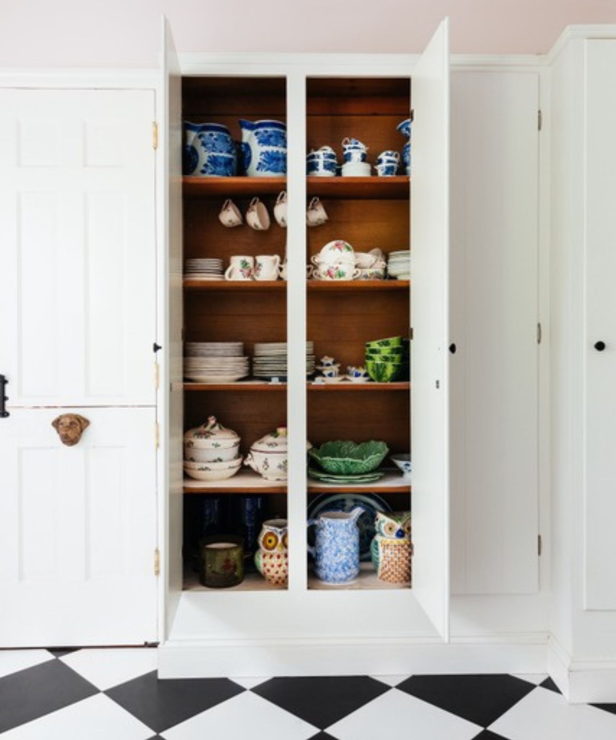 Built-in kitchen cupboard painted white with a pale pink ceiling, displaying colorful ceramics and tableware on warm wood shelves above a black-and-white checkered floor.