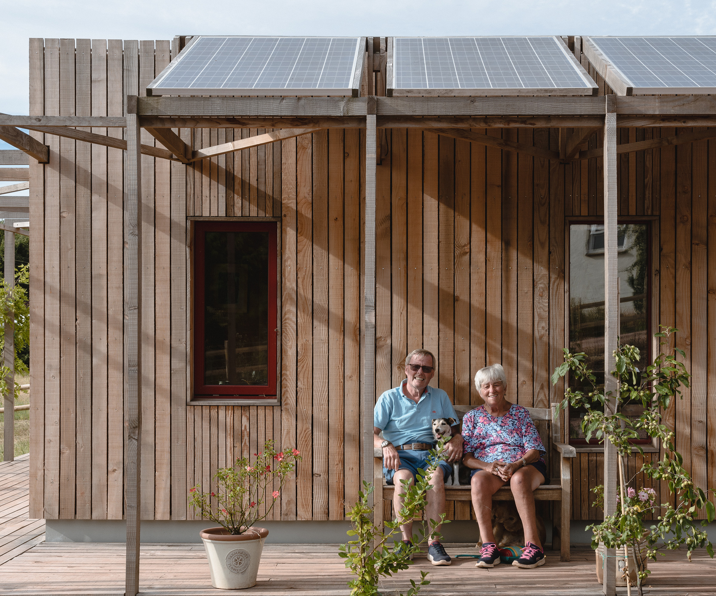 Couple sits in front of a timber clad single storey self build. There is a level access veranda with awning housing several solar panels