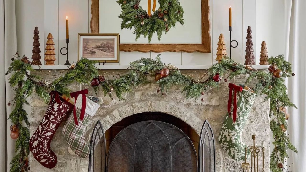 Stockings hanging from a garland above a fireplace; candles and Christmas tree figurines along a mantel against a white wall.