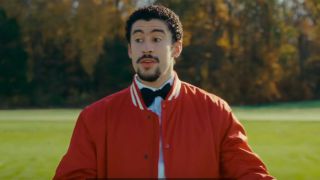 Bad Bunny looking like 'I dunno!' in a red jacket while standing on the golf greens in Happy Gilmore 2.