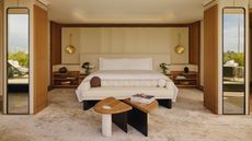Image of a luxury hotel bedroom. The bed is in the center with a white bench in front of it and an extended headboard with wooden side table attached. There is a coffee table in front of the bench and two columns in the room that have mirrors on them.