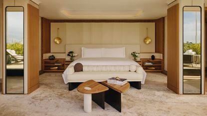 Image of a luxury hotel bedroom. The bed is in the center with a white bench in front of it and an extended headboard with wooden side table attached. There is a coffee table in front of the bench and two columns in the room that have mirrors on them.