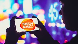 Bandai Namco logo displayed on a landscape-held smartphone with a city's neon lights in the background