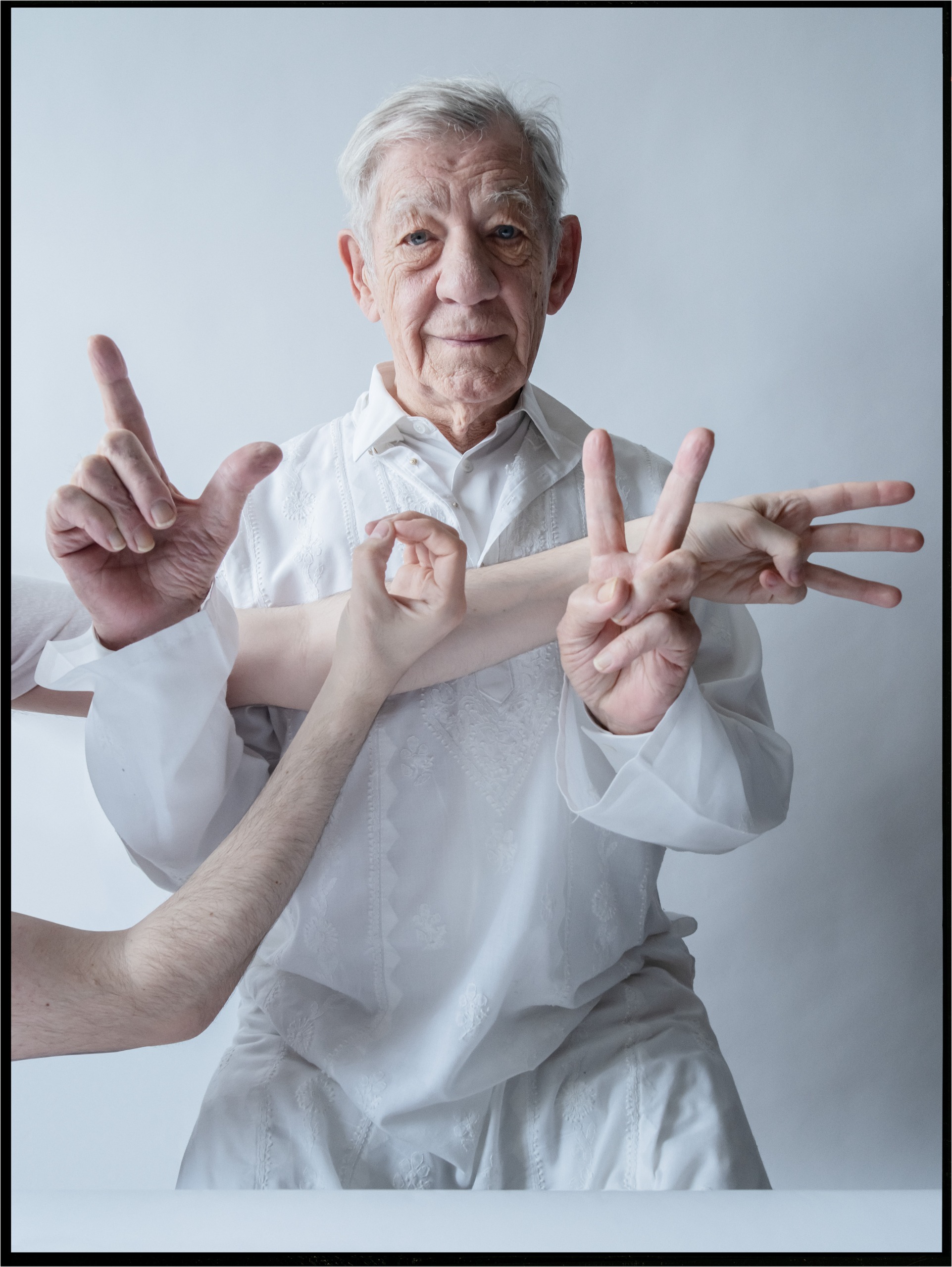 Photo of Ian McKellen in white embroidered clothing against a white background, with his hands and several other people's hands forming the letters of the word love in front of him.
