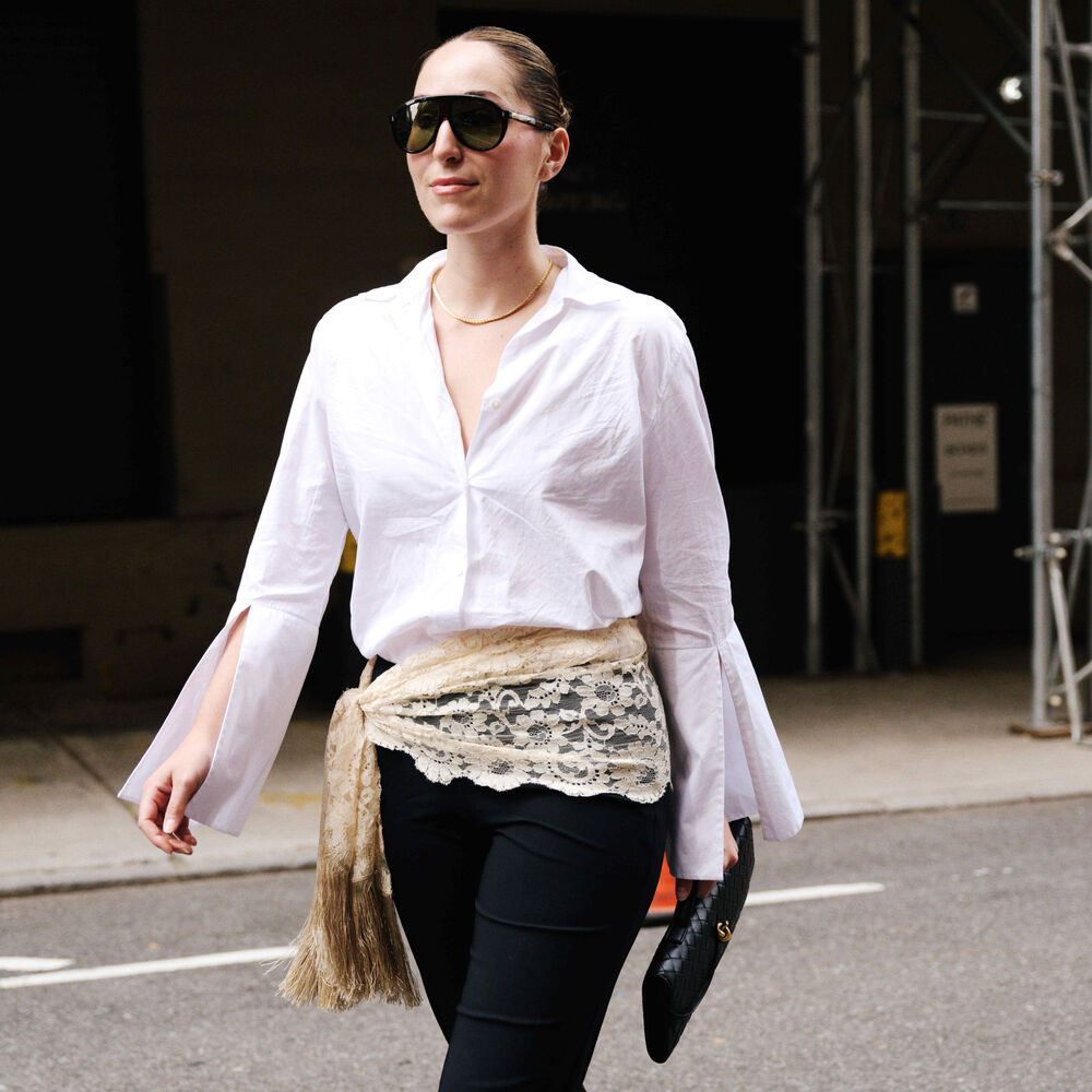 Anna wears white button down, black pants, and lace scarf tied around her hips. She is also carrying a bottega andiamo clutch.