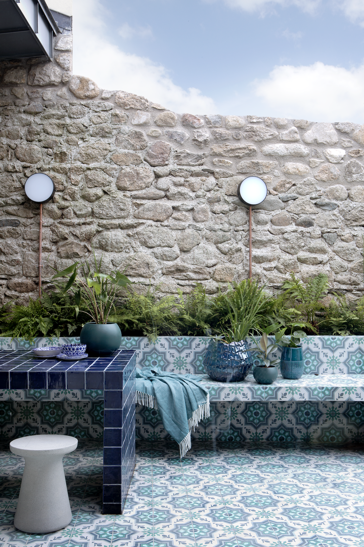 A patio area with blue pattern tiles on the floor and seating and a stone brick wall behind with wall lights and plants along the back of the seating with a gloss tile table