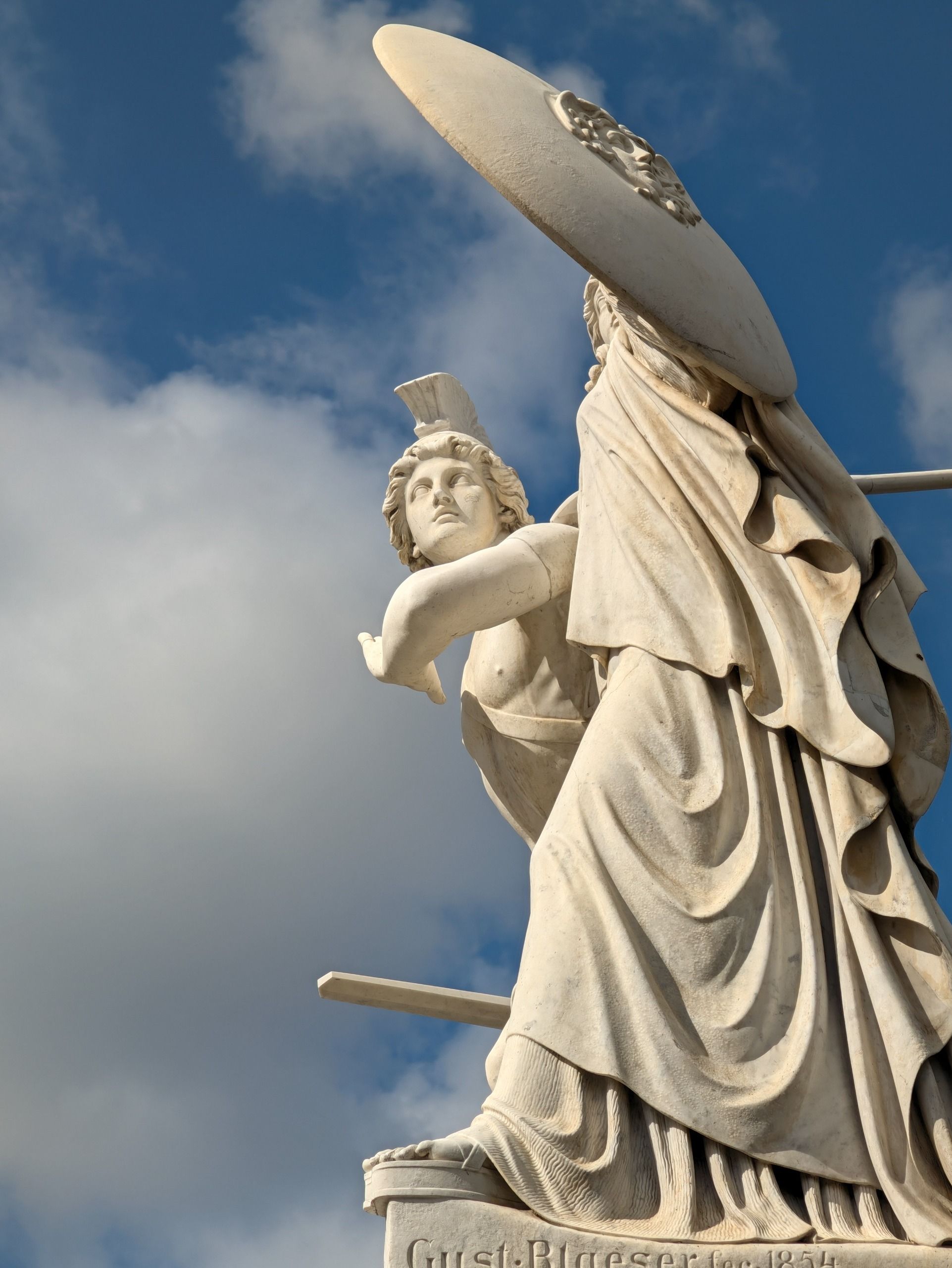 Low-angle view of a pale stone classical statue holding a large shield against a blue and cloudy sky.