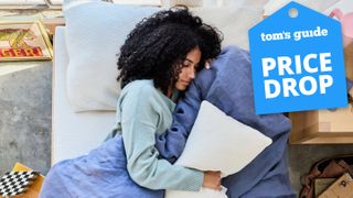 A college student lies on her side in bed, sleeping on the Avocado Eco Organic mattress topper with an Avocado Organic pillow beneath her head and one in her arms. She's wrapped in a blanket. A Tom's Guide Price Drop deals graphic in the top right corner