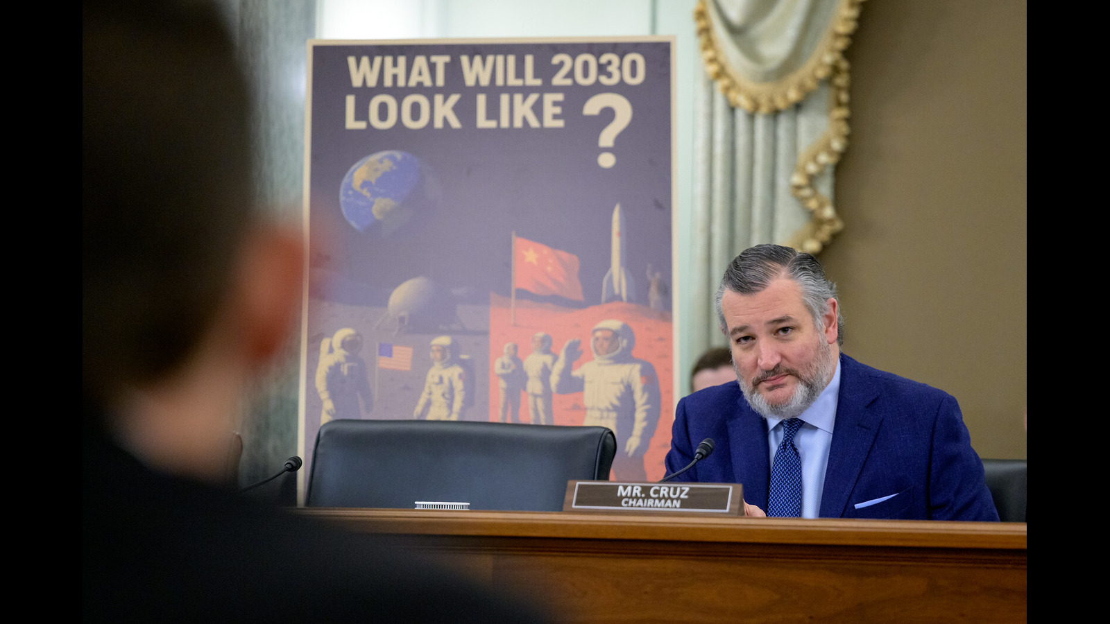 A man with a suit and tie sits behind a desk and looks at an audience off-camera