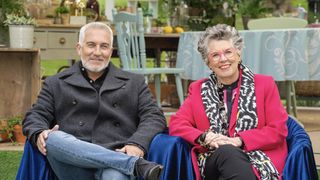 The Great Celebrity Bake Off 2023 — Paul Hollywood in a dark shirt and Prue Leith in a pink jacket and glasses sit next to each other in the tent in The Great British Bake Off.