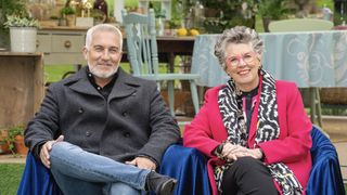 Paul Hollywood in a dark jacket and Prue Leith in a pink jacket and glasses sit next to each other in the tent in The Great British Bake Off.