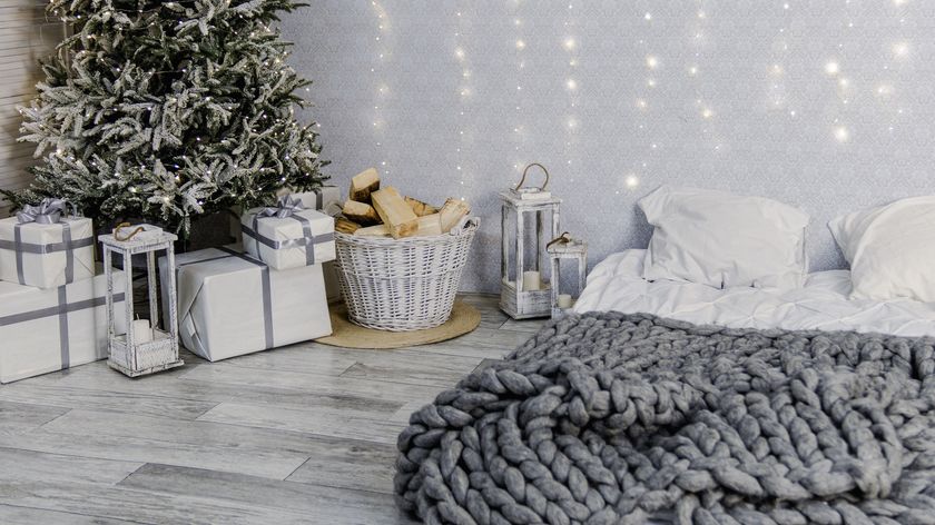 Image shows a spare room mattress placed on the floor and covered with white linens and pillows while a frosted green christmas tree sits in the corner surrounded by brightly wrapped presents 