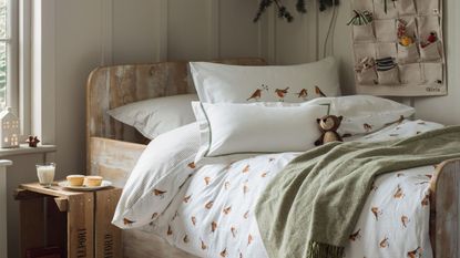 A white panneled bedroom with a wooden single bed, adorned with white robin bedding, a green throw blanketm and a small teddy. Mince pies and milk sit on a nightstand beside.
