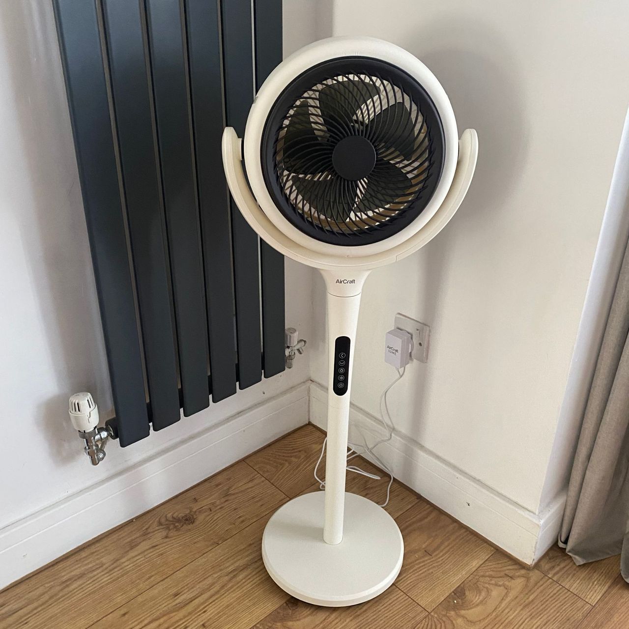 AirCraft LUME Quiet Air Circulator Fan in pedestal mode in the corner of a room on a wood-look floor next to a vertical radiator