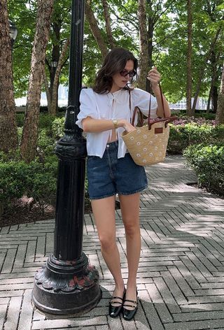 a woman's outfit showing what to wear in 80-degree weather with a white puff sleeve blouse styled with denim shorts and black mary jane ballet flats and a woven raffia tote bag