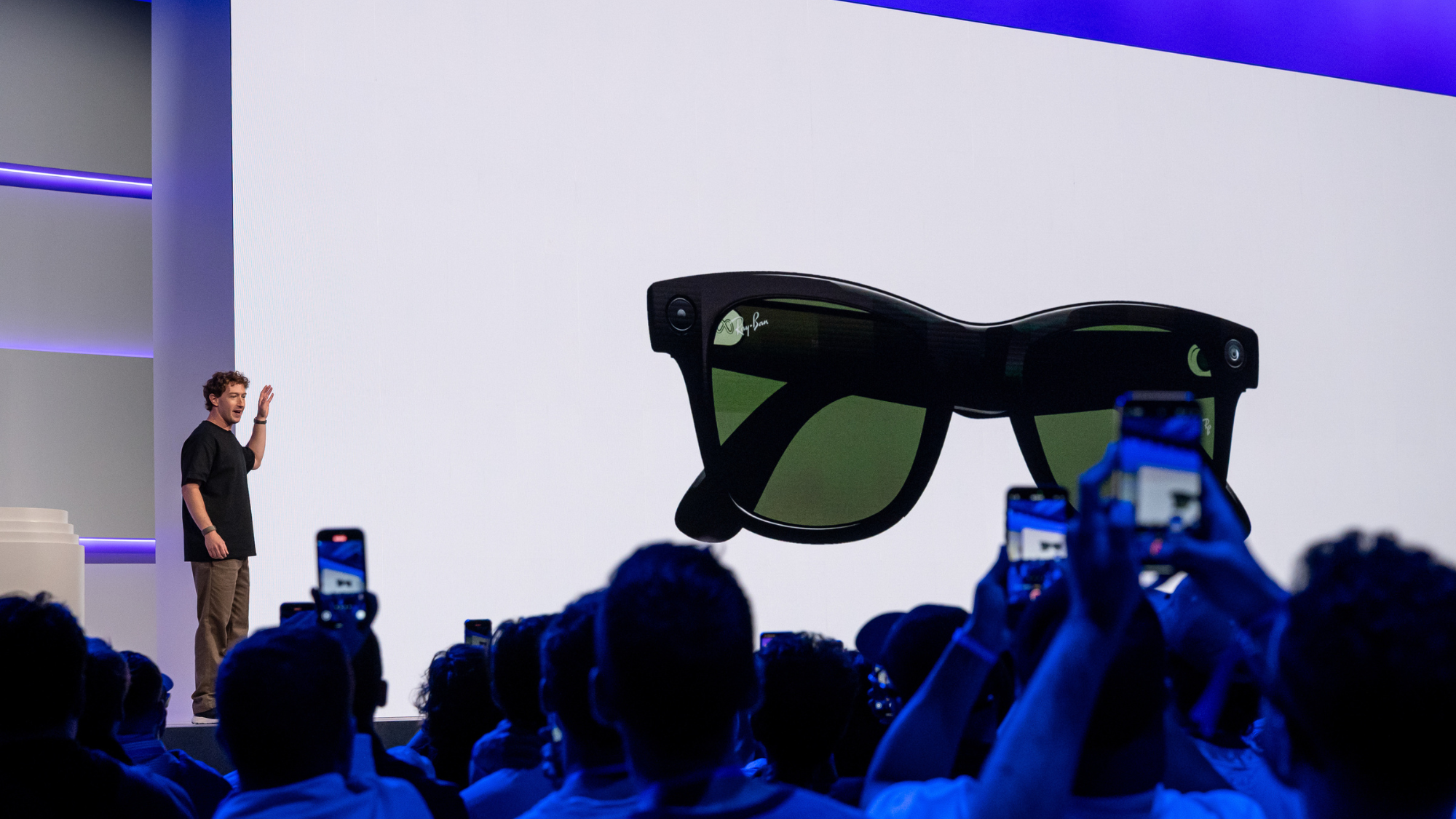 Mark Zuckerberg, wearing brown trousers and a black shirt, gestures at a large white screen showing a pair of black smart sunglasses